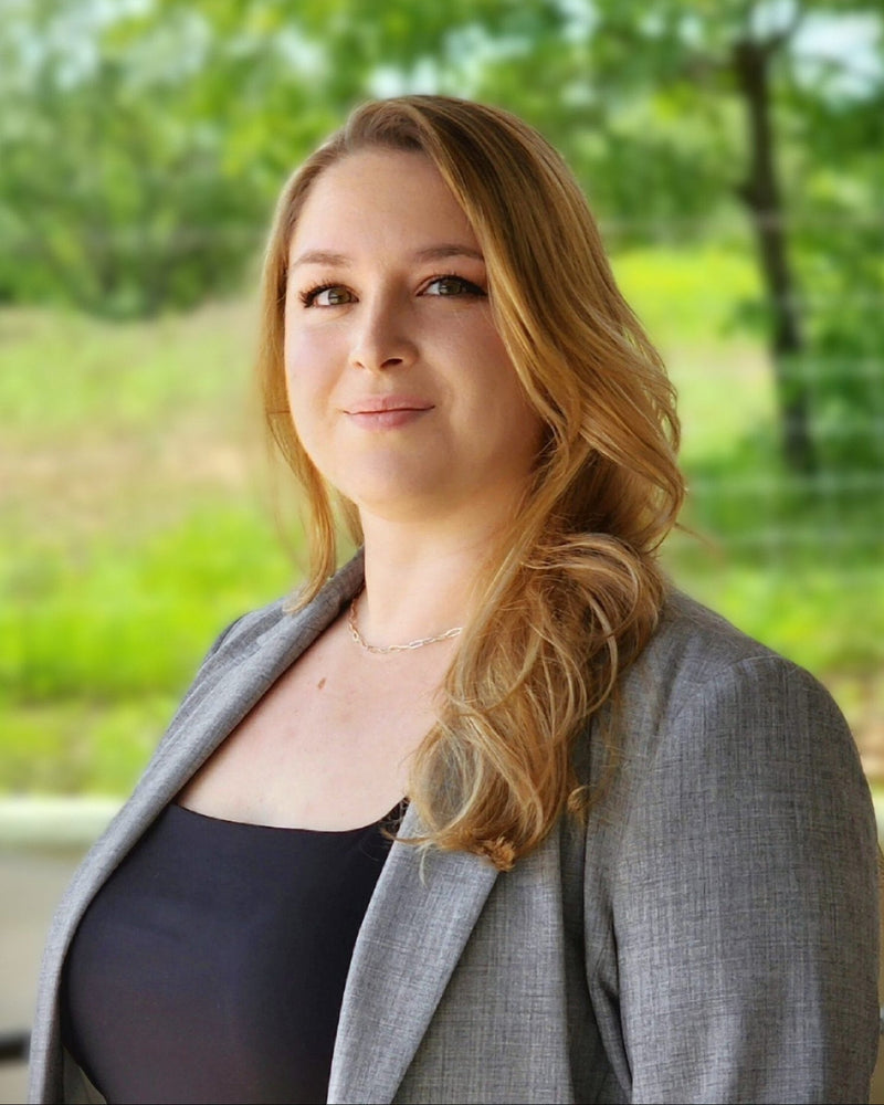 Samantha Dangerfield in a gray blazer standing outdoors with greenery in the background